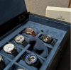A luxury watch display box with a blue velvet interior, showcasing several Omega wristwatches in individual slots. A white Omega-branded box is partially visible in the background.