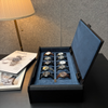 A black textured watch storage box with an open blue velvet interior holding ten watches, placed on a dark table next to a lamp and a magazine.