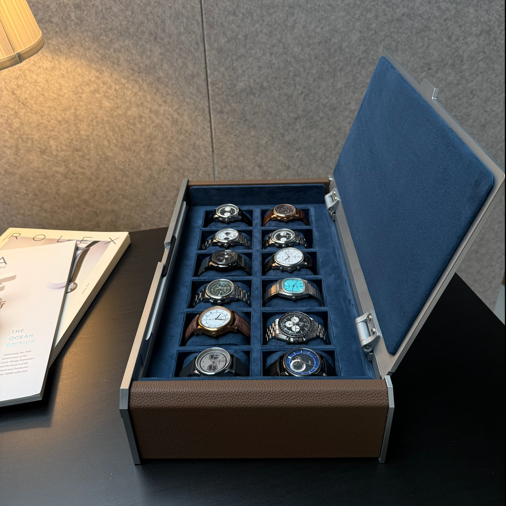 A luxury watch display box with an open lid, showcasing 12 different wristwatches arranged neatly inside. The box features a textured brown exterior, a deep blue velvet interior, and silver-tone metal hinges.