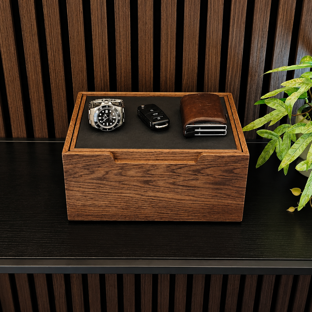 A rectangular wooden watch box with a dark interior tray holding a silver wristwatch, a black car key fob, and a brown leather wallet, displayed on a dark wooden shelf with a slatted wood wall background and a green plant at the side.